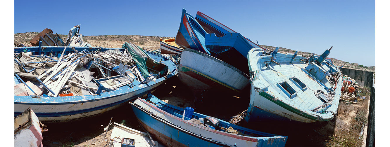 Small wrecked boats are piled atop one another on a desolate beach.