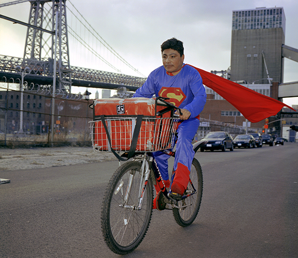A man in a Superman costume rides a delivery bike.